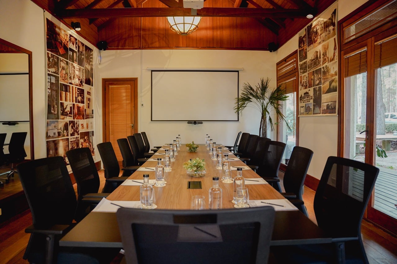 Salón Nomad: sala de reuniones con grandes ventanales, mesa larga con sillas y vegetación tropical al fondo.