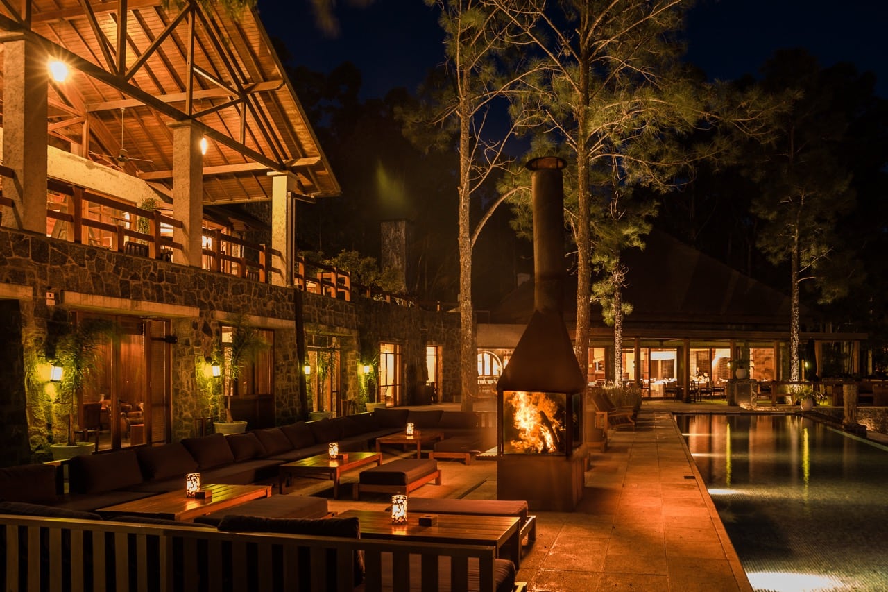 Terraza Mandara de noche: hogar a fuego, sillones bajos y piscina iluminada bajo los pinos.