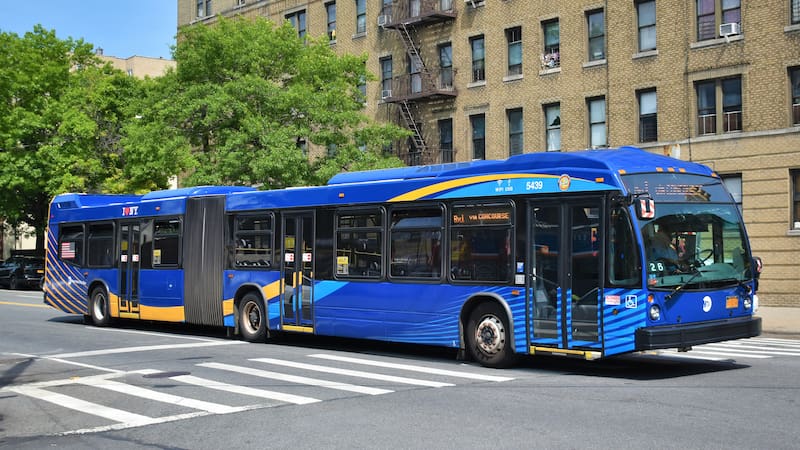 A driver of an MTA New York City Transit Nova Bus LFS articulated bus, 5439, is preparing to turn left from Bailey Avenue onto Albany Crescent, while operating along the Bx1 Limited in Kingsbridge, due south to Mott Haven.