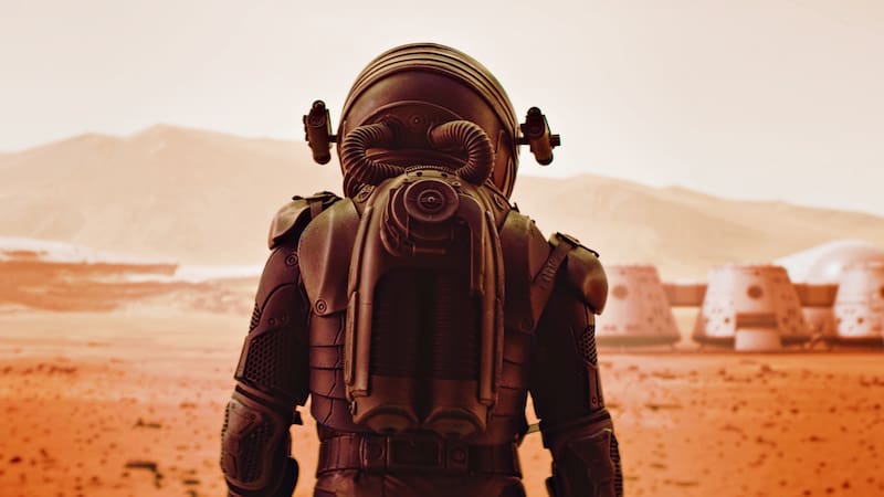 Back view of astronaut wearing space suit walking on a surface of a red planet. Martian base and rover in the background. Mars colonization concept