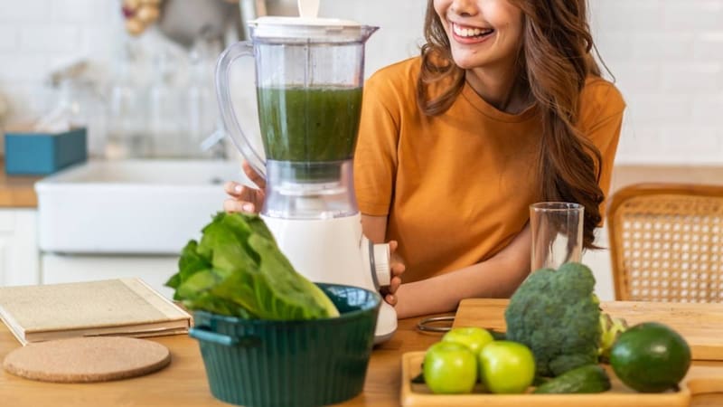 Mujer prepara un batido verde de frutas y verduras en una cocina.