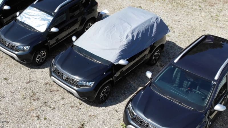 Una vista aérea de varios vehículos estacionados al aire libre, donde se aprecian diferentes accesorios de protección solar, destacando una lona cubriendo la parte superior de un SUV negro.