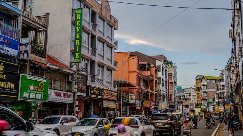 Cars and motorbikes travel along a busy street lined with multi-story buildings and colorful shop signs in Da Lat. A green vertical sign for Hotel Reverie is visible on one of the facades. Da Lat, Vietnam - May 19, 2023