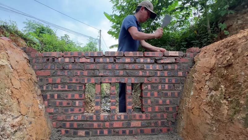 Un constructor levanta manualmente un muro de contención de ladrillos y cemento en el cauce de un arroyo, incorporando compuertas centrales para regular el flujo de agua.