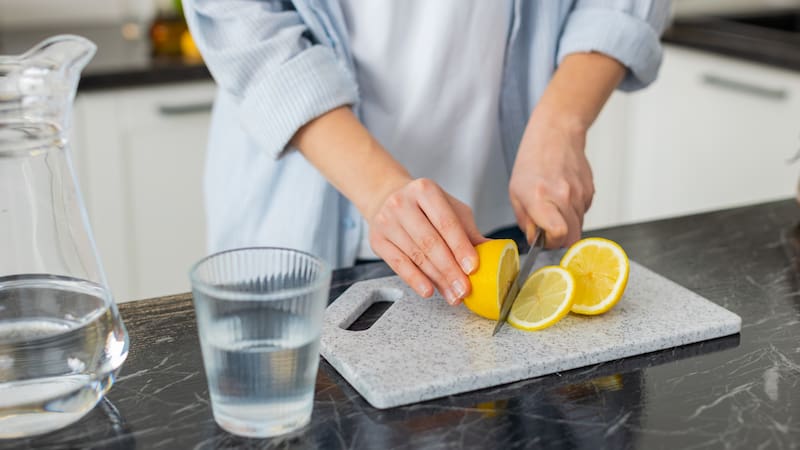 Hands cutting fresh lemon on board in kitchen to make vitamin water for hydration and detox. Close-up food prep with knife glass, carafe for fit morning drink and clean eating simple daily routine