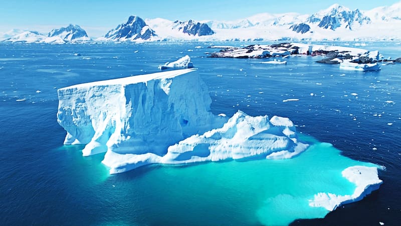 Melting iceberg in Antarctica in sunny day. Huge glacier in turquoise sea water, snow covered mountains in background. Aerial drone flight panorama. Global warming, climate change. Explore South Pole