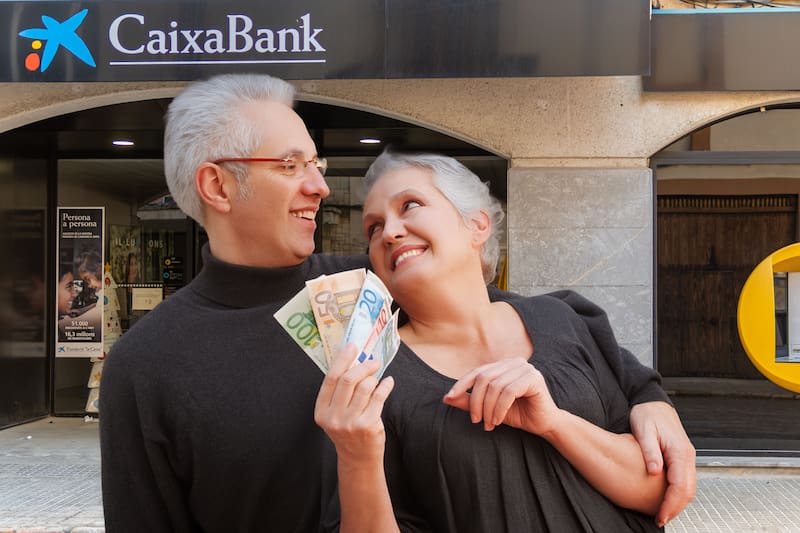 Pareja de adultos mayores frente a una oficina de CaixaBank con euros en la mano.