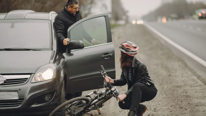 Una ciclista con casco y chamarra de cuero permanece agachada junto a su bicicleta caída tras un percance, mientras un conductor sostiene la puerta abierta de su vehículo gris e interactúa con ella a la orilla de la carretera.