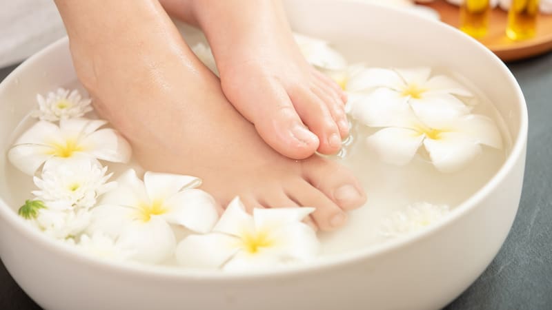 closeup view of woman soaking her feet in dish with water and flowers on wooden floor. Spa treatment and product for female feet and hand spa. white flowers in ceramic bowl.