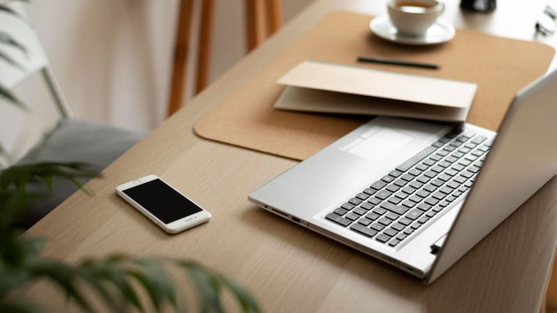 Una mesa de trabajo iluminada que cuenta con una computadora portátil abierta, un teléfono inteligente, un cuaderno de notas y una taza de café en el fondo.