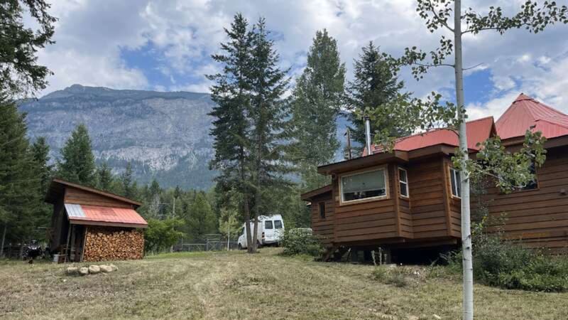 Casa de madera en el bosque con un depósito de leña apilada y montañas rocosas de fondo.